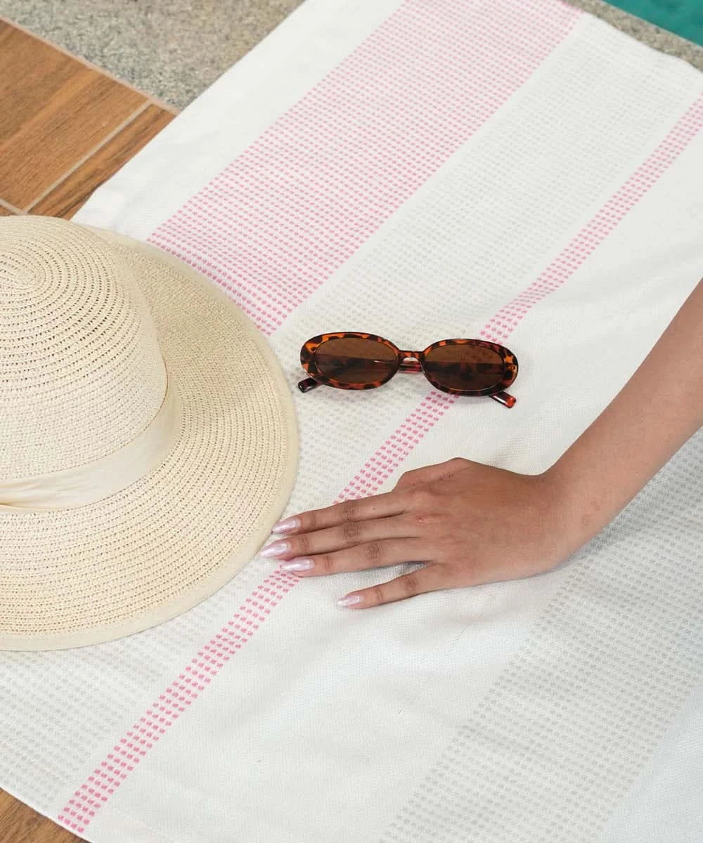 Beige straw hat and tortoiseshell sunglasses on a white and pink striped towel with a hand reaching out.