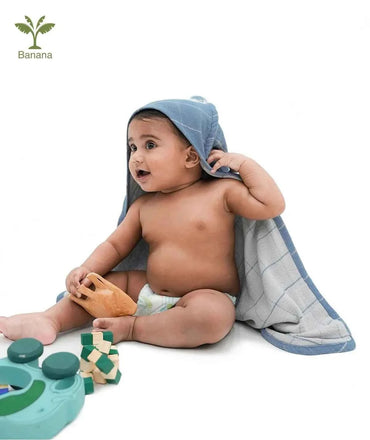 Baby sitting on a white surface with a gray towel draped over its head, surrounded by toys.