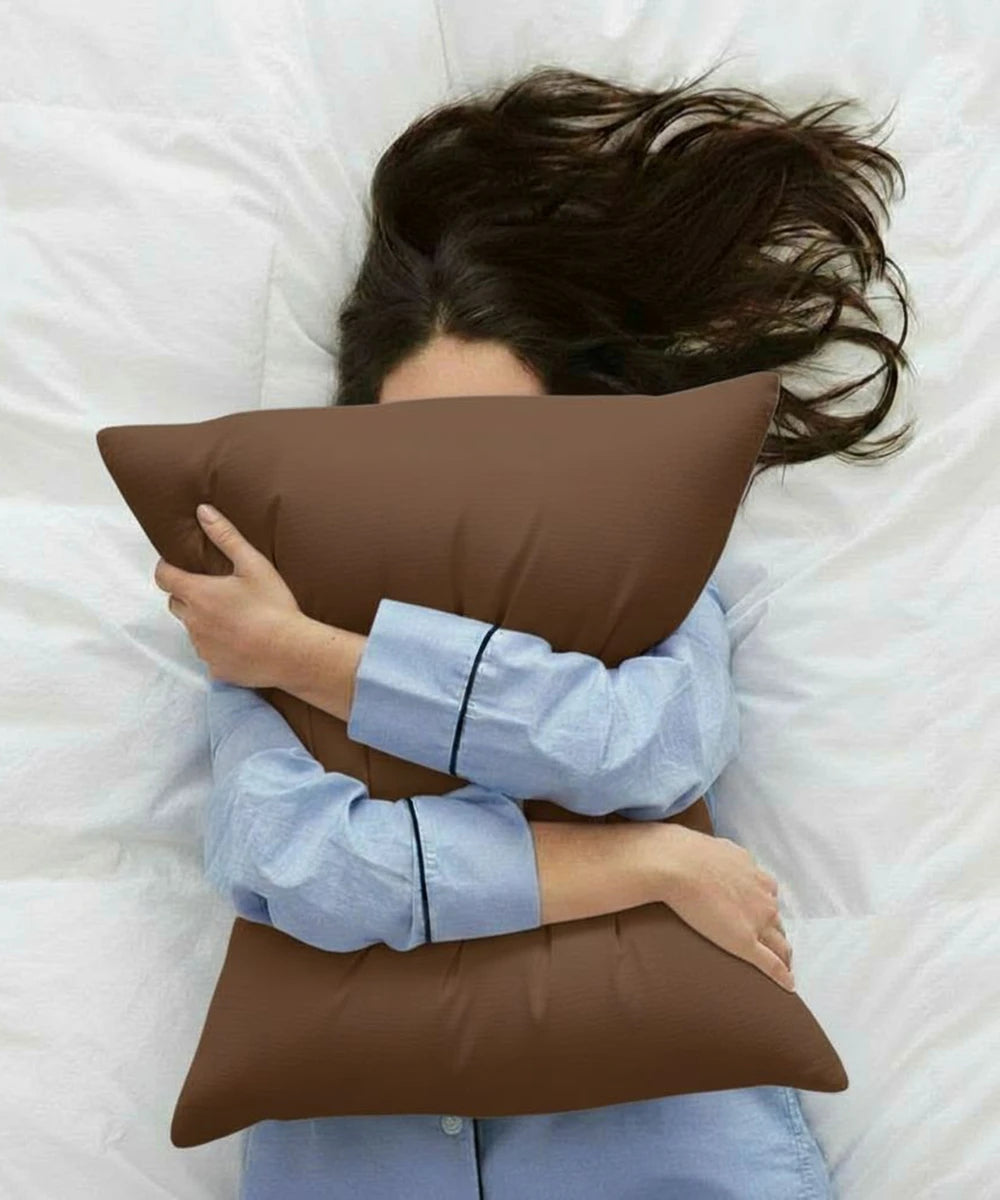 Person in blue pajamas lying on a white bed, hugging a chocolate-brown pillow with a House of Doctor pillowcase.