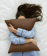Person in blue pajamas lying on a white bed, hugging a chocolate-brown pillow with a House of Doctor pillowcase.