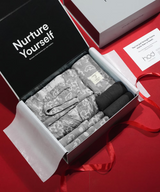 Open gift box with gray lace lingerie on a red surface, featuring 'Nurture Yourself' text.