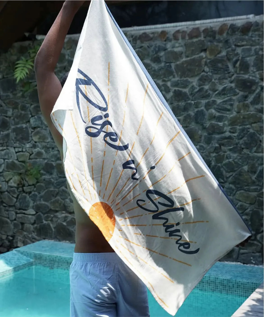Person holding a towel with 'Rise & Shine' text by a poolside.