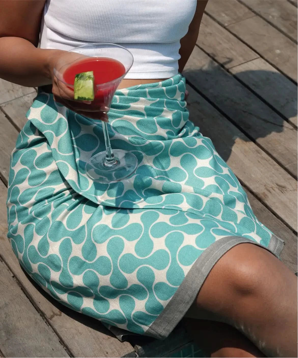 Person wearing a teal and white patterned skirt holding a cocktail on a wooden deck.