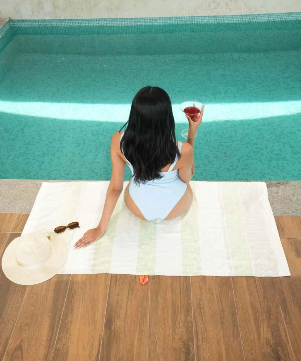 Woman in a blue swimsuit sitting on a bamboo dobby towel by a pool holding a drink.
