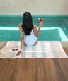 Woman in a light blue swimsuit sitting by a pool holding a drink, with a bamboo dobby towel and hat on the floor.