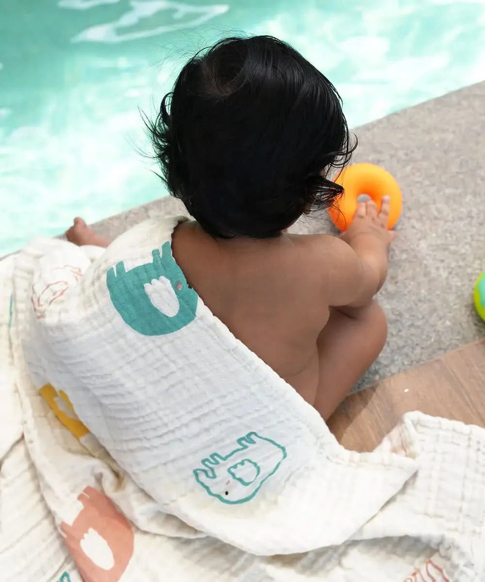 Baby sitting on a towel by a pool holding an orange toy.
